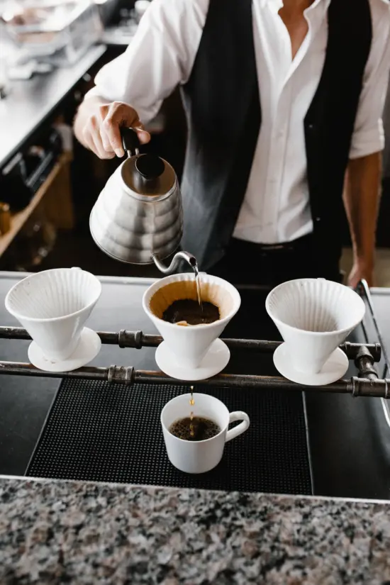 A barista in a white shirt and vest pours water from a gooseneck kettle over a pourover dripper. Three drippers are placed over two horizontal bars creating a stand, with room to put mugs underneath.