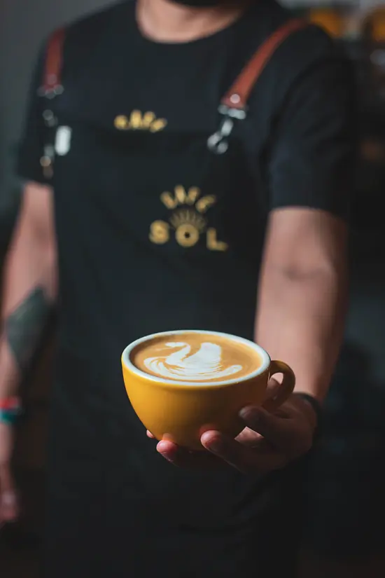 A barista in a black cafe sol apron holds out a yellow mug with swan latte art.