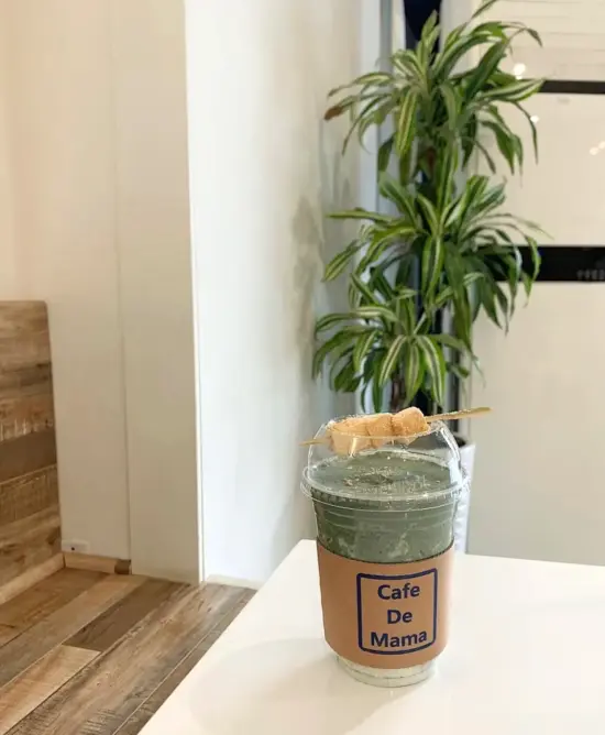 A green cold drink with small pieces of rice cake skwered and set on top of the plastic lid. The cup has a paper sleeve labeled Cafe de Mama. There is a potted plant 