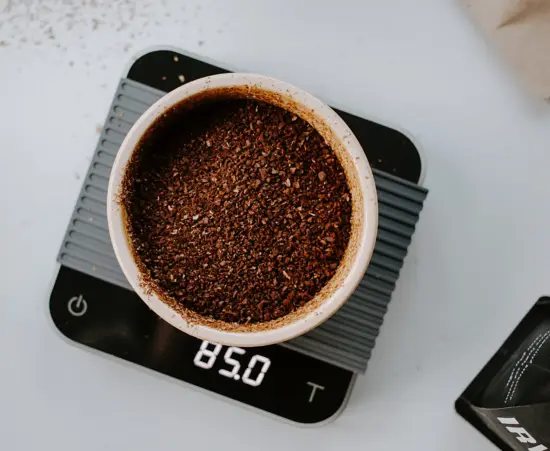 A black scale with a gray mat holds a white ceramic cup with course coffee grounds on top of a white surface.