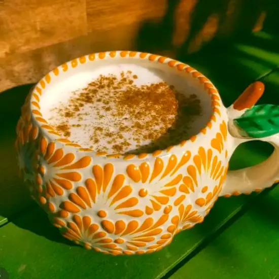 Una taza hecha a mano con flores amarillas en relieve y un patrón de hojas en el asa sostiene una bebida caliente espolvoreada con canela.