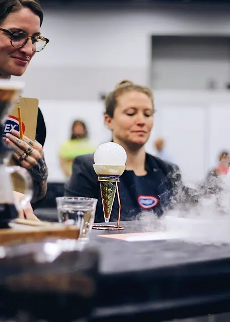 Two judges look at Sam Schroeder's cold cocktail, which looks almost like an ice cream cone, having a large round bubble on top and a rainbow colored cone shaped cup on a stand.