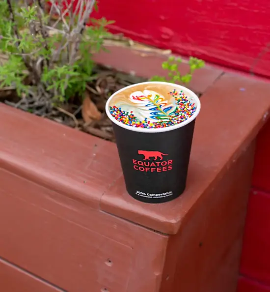 An Equator paper cup with a latte featuring rainbow milk swirls and round rainbow sprinkles on top.