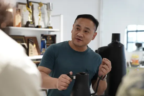 Jingui Zhu holds a kettle and talks to customers in his cafe and he makes coffee.