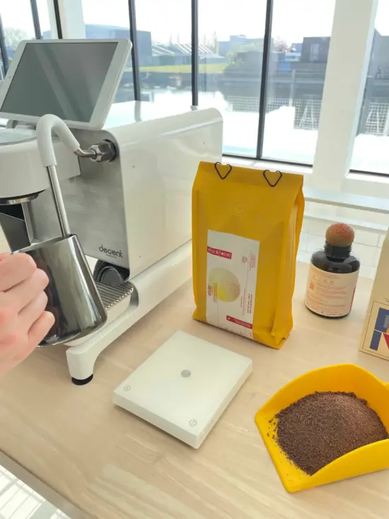 A steam wand, scale, bag of cacao, and yellow scoop with cacao powder. A barista is using the steam wand and a metal pitcher.