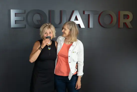 Two women hug for a photo, one drinking from a black paper coffee cup and wearing a heart shaped pride flag necklace.