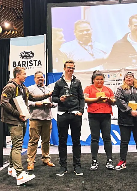 Sam Schroeder stands with other competitors at USCIGS. He holds a glass rectanglular trophy, wears a gray jacket and jeans, has glasses and short hair.