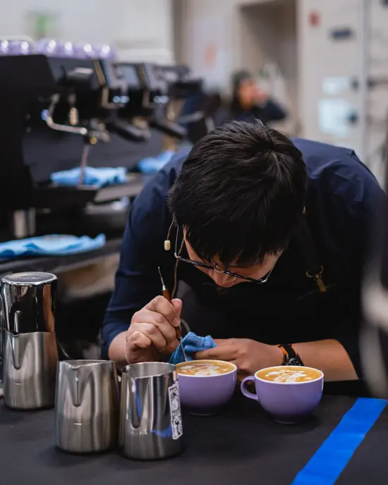 Flook leans over two lattes with a thin tool, adjusting his designs.