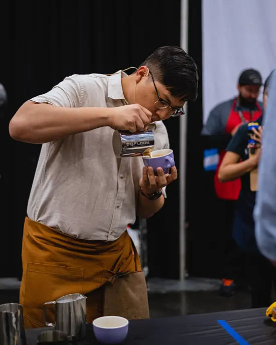 Flook in an off white shirt, glasses, and tan apron pours latte art in a purple mug. A judge with a timer watches behind the competition table.