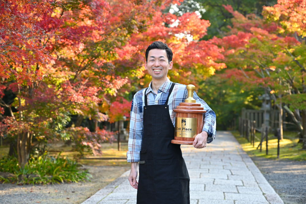 Japanese barista champion Toshio Uchibe poses with his first-place trophy with trees in autumn colors behind him.