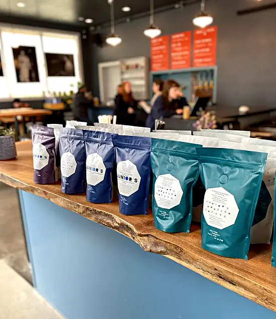 A natural wood counter holds blue, white and purple coffee bags with the Juniors logo. People are seated in the background at cafe tables.