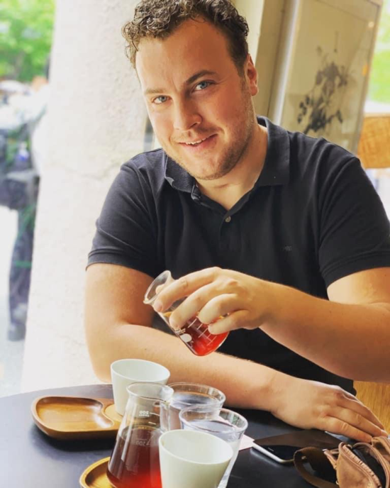 Niels te Vaanhold pours some coffee into a white ceramic cup from a small glass container as he smiles at the camera.