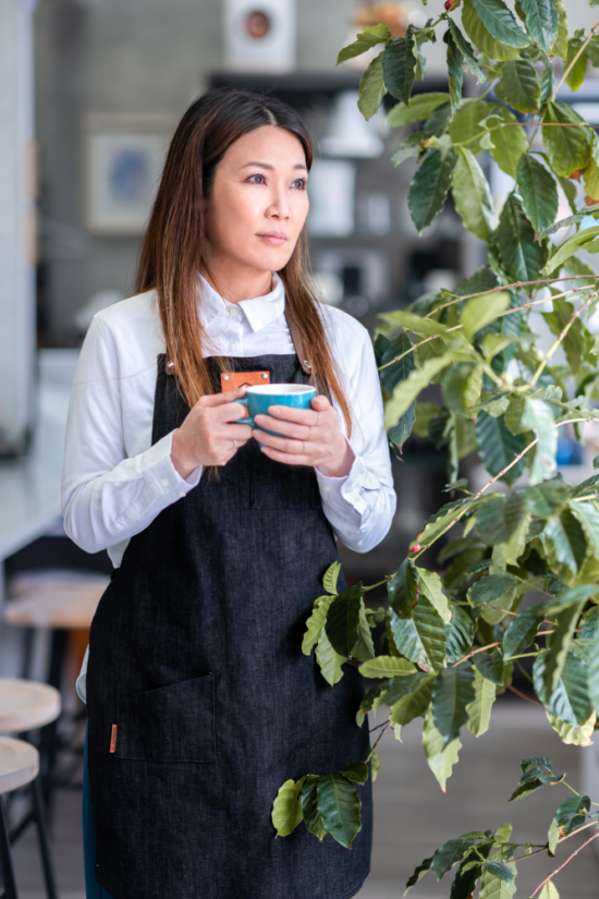 Hanna Teramoto holds a turquoise ceramic coffee cup in both hands as she stands next to a coffee plant inside of a cafe.