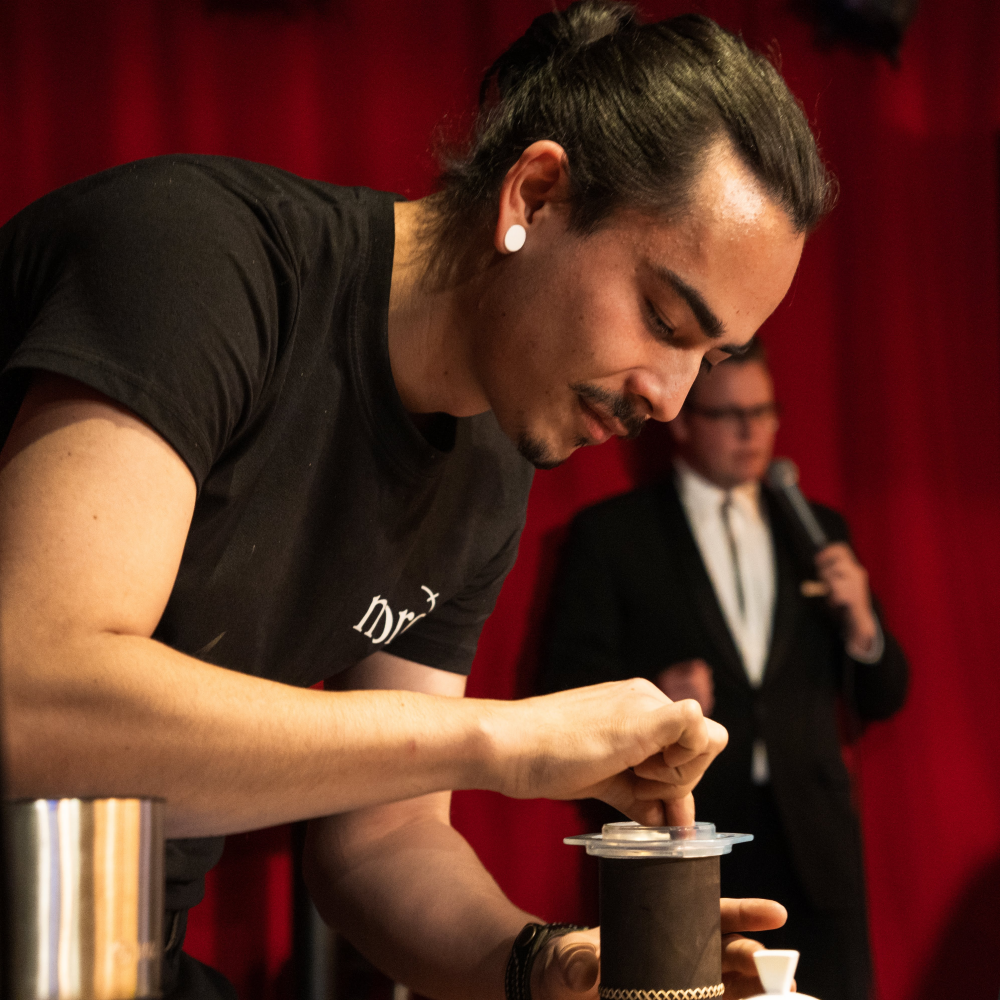 Alexander Monsen prepares a coffee during a competition to earn a place at the World Barista Championship while an emcee speaks behind him.