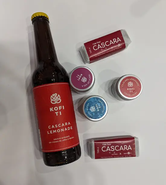 Kofi Ti's cascara lemonade is packaged in amber longneck bottles with a red label. Also in the photo are two small cascara snack bars and three small metal tins.