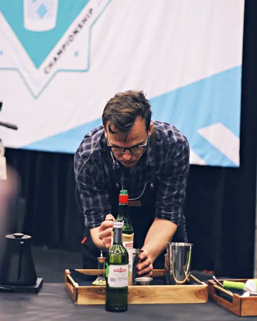 Sam Schoeder works on one of his signature drinks during the competition. there are two liquor bottles on the table, some wooden trays, a jigger and Boston style cocktail shaker.