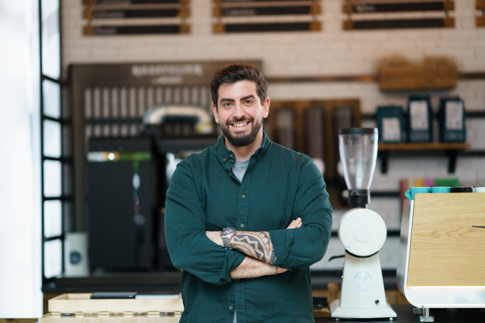 Chris Sotiros poses in front of a table with his arms crossed and smiles at the camera. On the table behind him are an espresso machine and a grinder.
