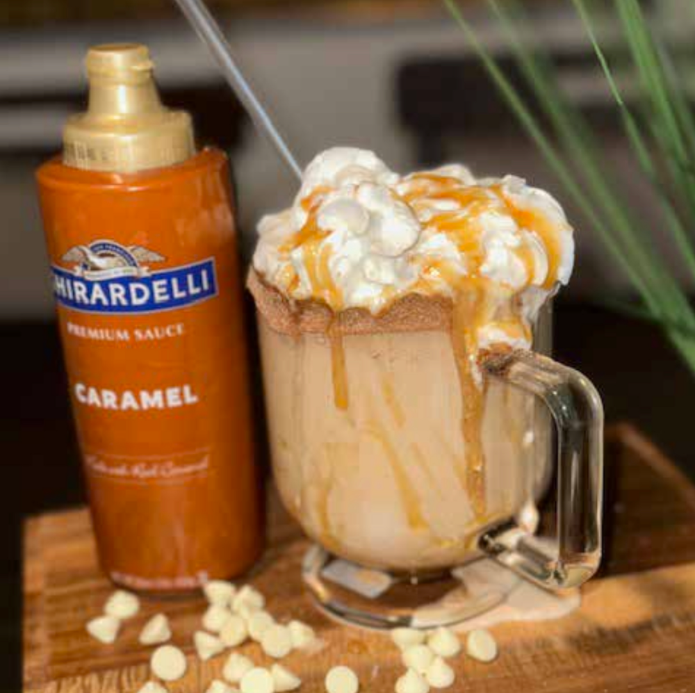 Tabitha Clarke's Cinnamon Roll in a Cup creation for the Ghirardelli Cool Caramel Challenge has whipped cream and caramel overflowing the top of a glass next to a bottle of caramel sauce.