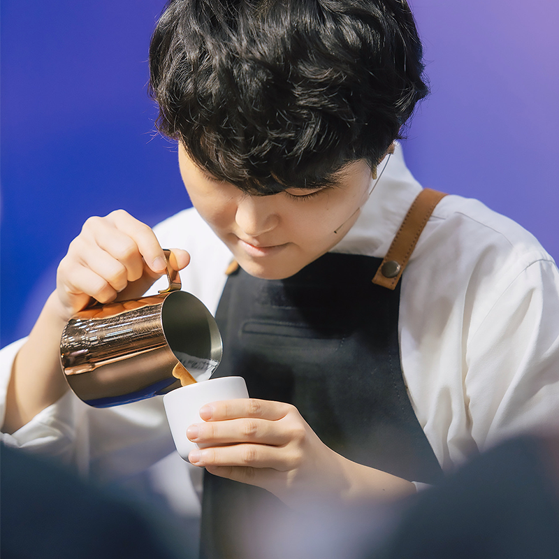 Jane Veraworawet pours steamed milk from a pitcher into a white ceramic cup to create latte art.