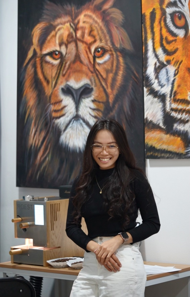 Mariam Erin poses in front of a table with an espresso machine on it. Behind her are paintings of a lion and a tiger.