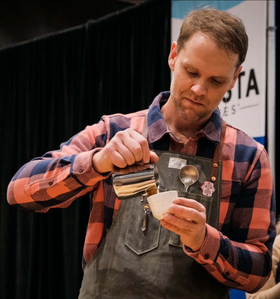 Isaiah Sheese pours milk from a steaming pitcher into a white ceramic cup during a barista competition. He is wearing a plaid shirt and canvas apron.