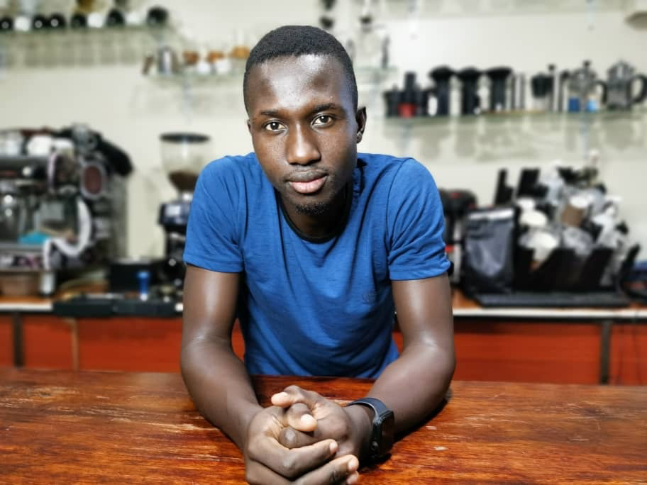 Wokorach Micheal leans forward on a bar with his hands clasped in front of him. Behind him is an espresso machine and grinder with other coffee equipment.