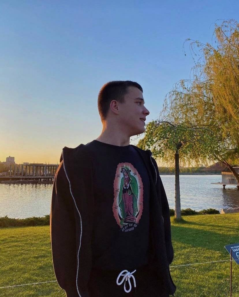 Ruslan Demonenko wears a black t-shirt and hoodie while standing in front on a body of water with some trees under a blue sky.