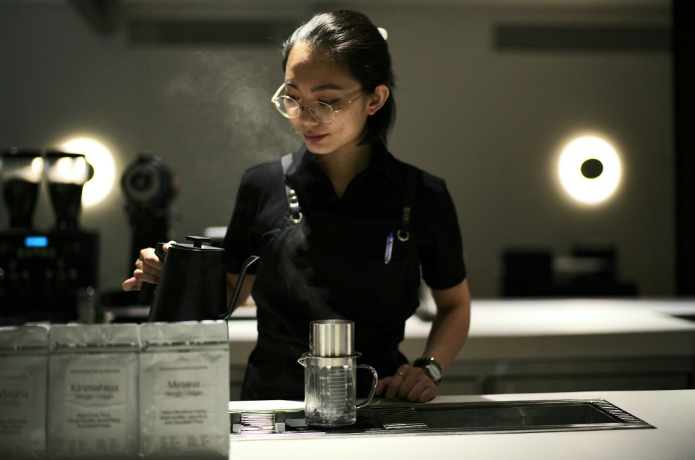 Han Tran holds a steaming kettle as she prepares to create a pourover in a dramatically lit cafe space.