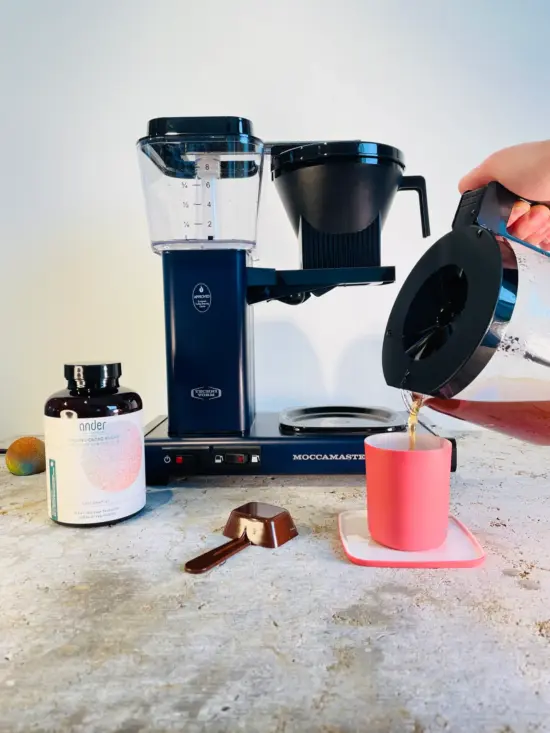 A Moccamaster, ander® bottle, and a pink mug with cacao drink being poured into it from a glass server.