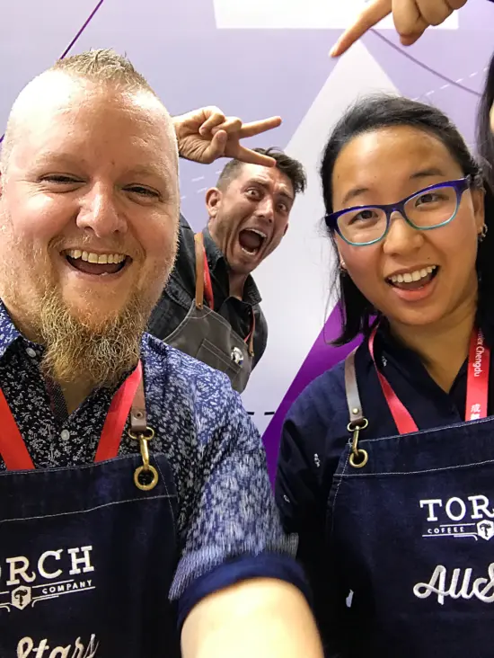 David poses with a woman in glasses and a man giving a peace sign. All three wear judge's aprons.