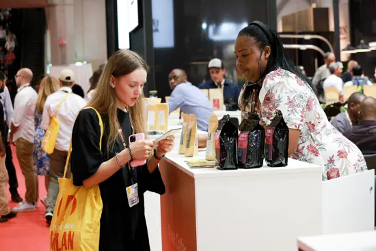 Two women at a counter on the show floor discuss coffee.