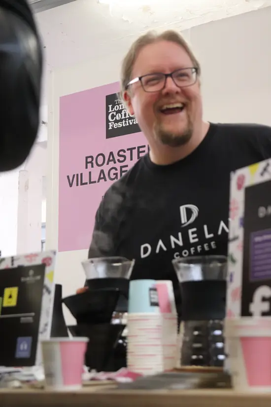 David at the Roasters Village in a coffee festival, wearing a Danelaw Coffee tee shirt. He has brewing devices set up on a table in front og him, blurred out of the photo.