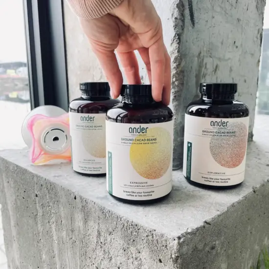 A hand places an ander® bottle next to two others on a concrete block. The bottles contain ground cacao. The circles on front of the bottles are different colors based on region and flavor profile.