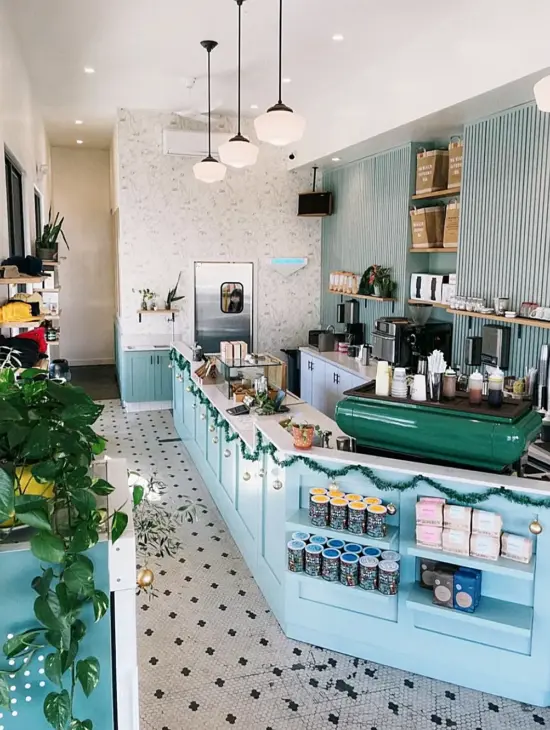 Interior of Steelhead with black and white tile floors, a light blue countertop, vibrant green espresso machine and vintage light fixtures. Greenery is sprinkled throughout. In-set wood shelves hold coffee bags on a textured sea green wall behind the counter.