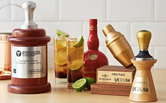 Several trophies and coffee cocktails are assmbled together on a counter. There is the large thimble-shaped World Coffee in Good Spirits award with a metal tamp handle on top and a wooden barrel shape on bottom, with a silver metal printed name and date.
There is a first place award for another compettition that has a plain wooden base and cocktail shaker in gold leaning across the top. There is also a tamper-shaped award on top of a pedestal that says Champion UK 2014.