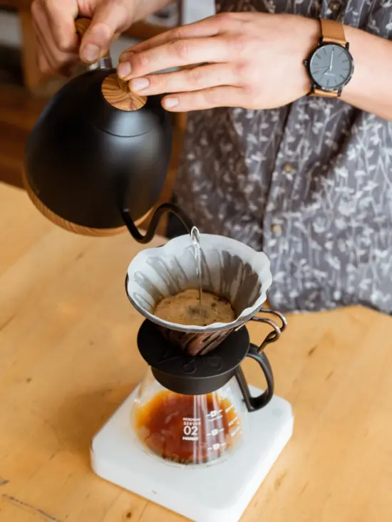 A pourover set up on a wooden table. The carafe sits on a white scale. Water is being poured from a black gooseneck kettle. 