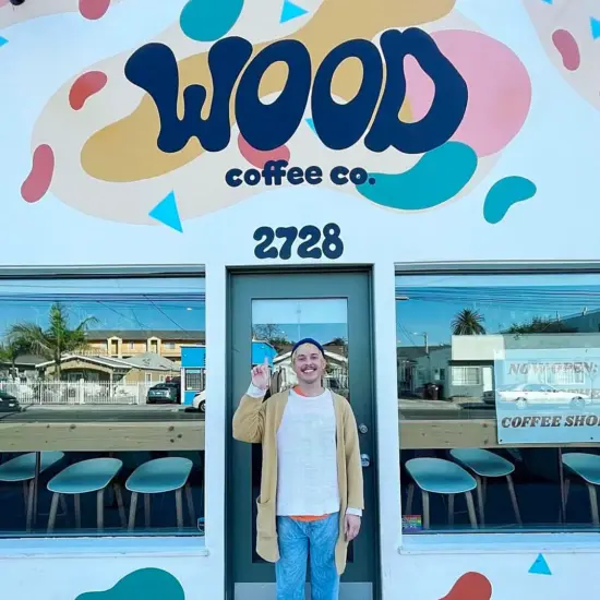 The storefront of Wood Coffee Co. with funky swirled paint patterns on the building, and bubble letters of the brand name above the door. Inside big windows is counter seating with minimalist white stools.