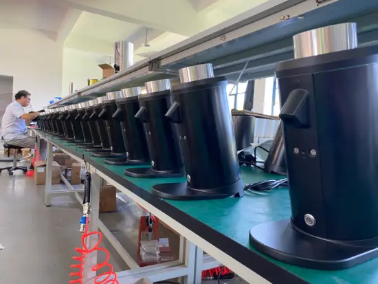 A man at one end of a long table works to assemble or calibrate a coffee grinder. there are over a dozen grinders lined up in a row waiting their turn.