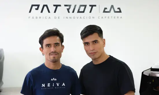 Two men pose for a photo in front of the Patriot logo on a white wall. 