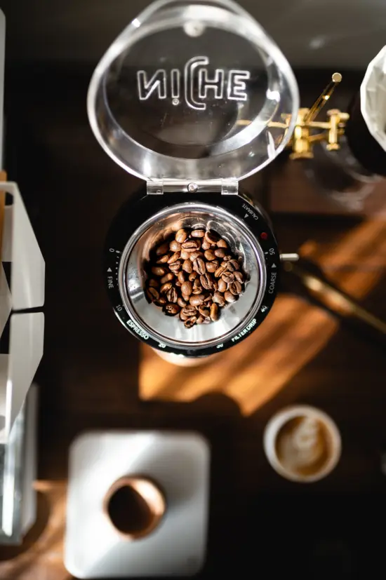 A black Niche grinder with dial and flip-top clear lid. Blurred out in the photo are a latte and a scale with a cup.