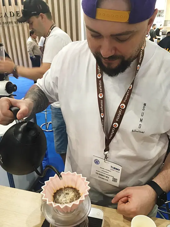 Daniel wears a backwards ball call and lanyard while brewing through a ceramic pourover.