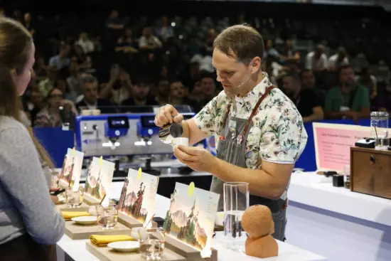 Isaiah pours milk during the finals, wearing a floral patterned shirt and denim apron. Judges watch in front of his table, and the audience watches from the background.