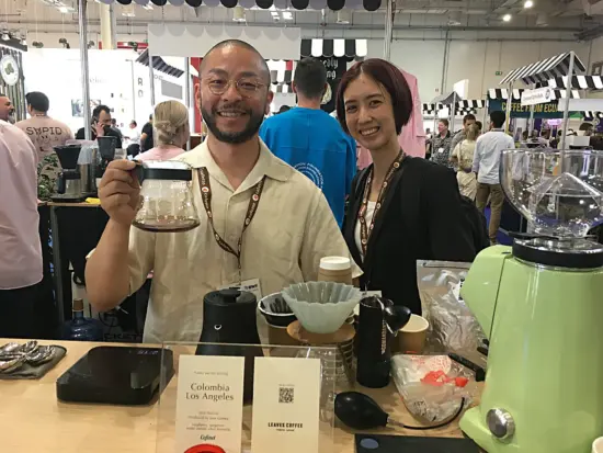 A man in glasses and woman in a blazer pose with their beans and gear, featuring a Colombian coffee.