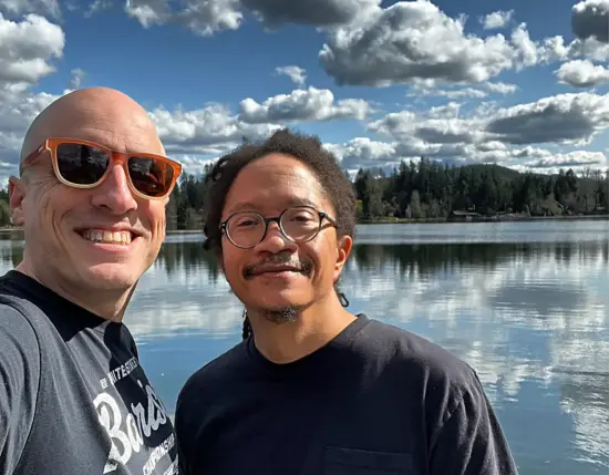 Nathanael y Lem posan frente a un lago cristalino que refleja el cielo azul y parcialmente nublado.