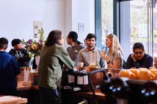 John talks to a man and woman at the caffe from behind the bar. other guests are crowded around the counter.