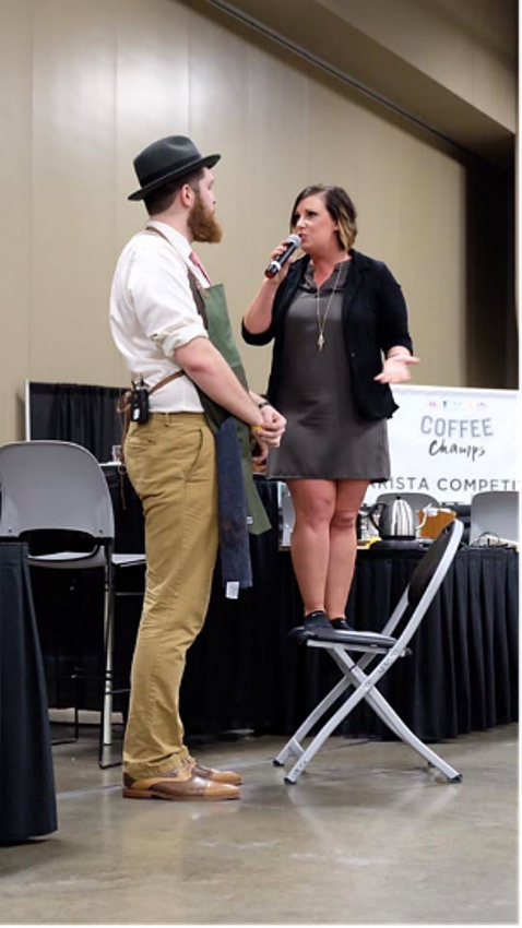 Anna stands on a chair with a microphone to be at eye level with a tall competitor she is talking to.