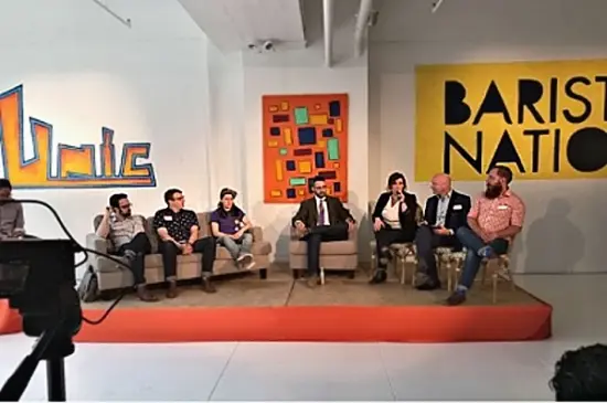 Anna and six others are seated on a small stage with a big Barista Nation sign behind them. Anna holds the microphone.