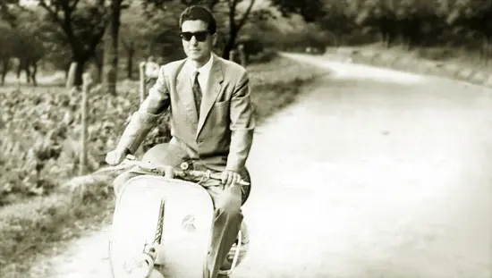 A young man on a motor scooter zips down a road in Italy. He wears a suit and tie and sunglasses.