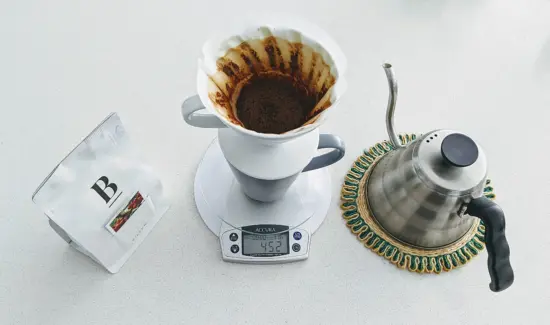 A white pourover setup with a gooseneck kettle, white scale, and bag of coffee beans.