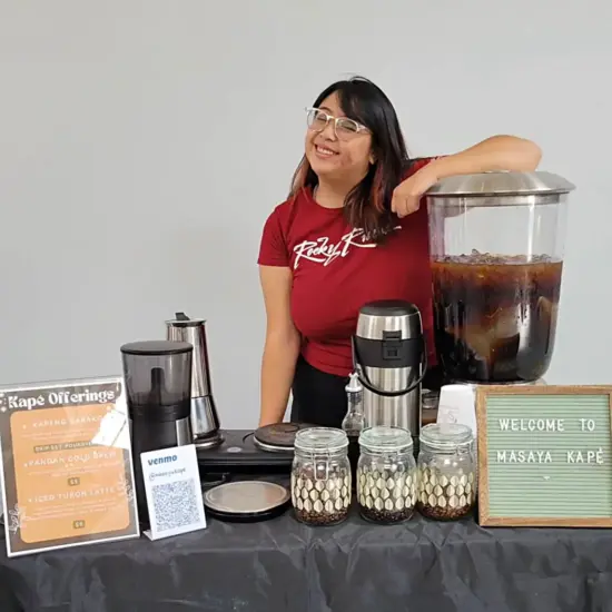 A barista at a coffee stand with cold brew, glass jars, a hot coffee pot and felt sign.